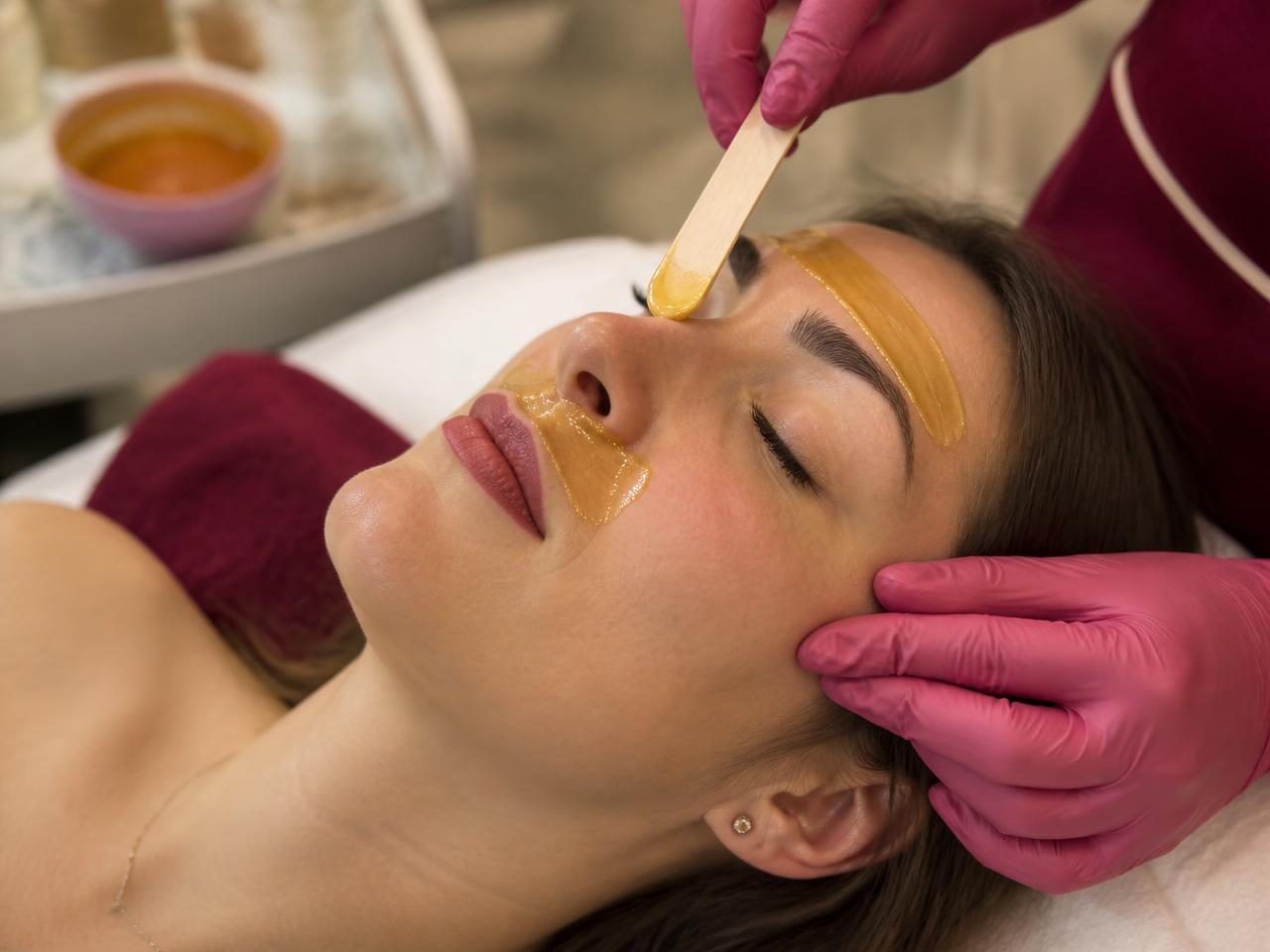 Facial waxing course showing warm wax applied to a client's brow and upper lip with a spatula.